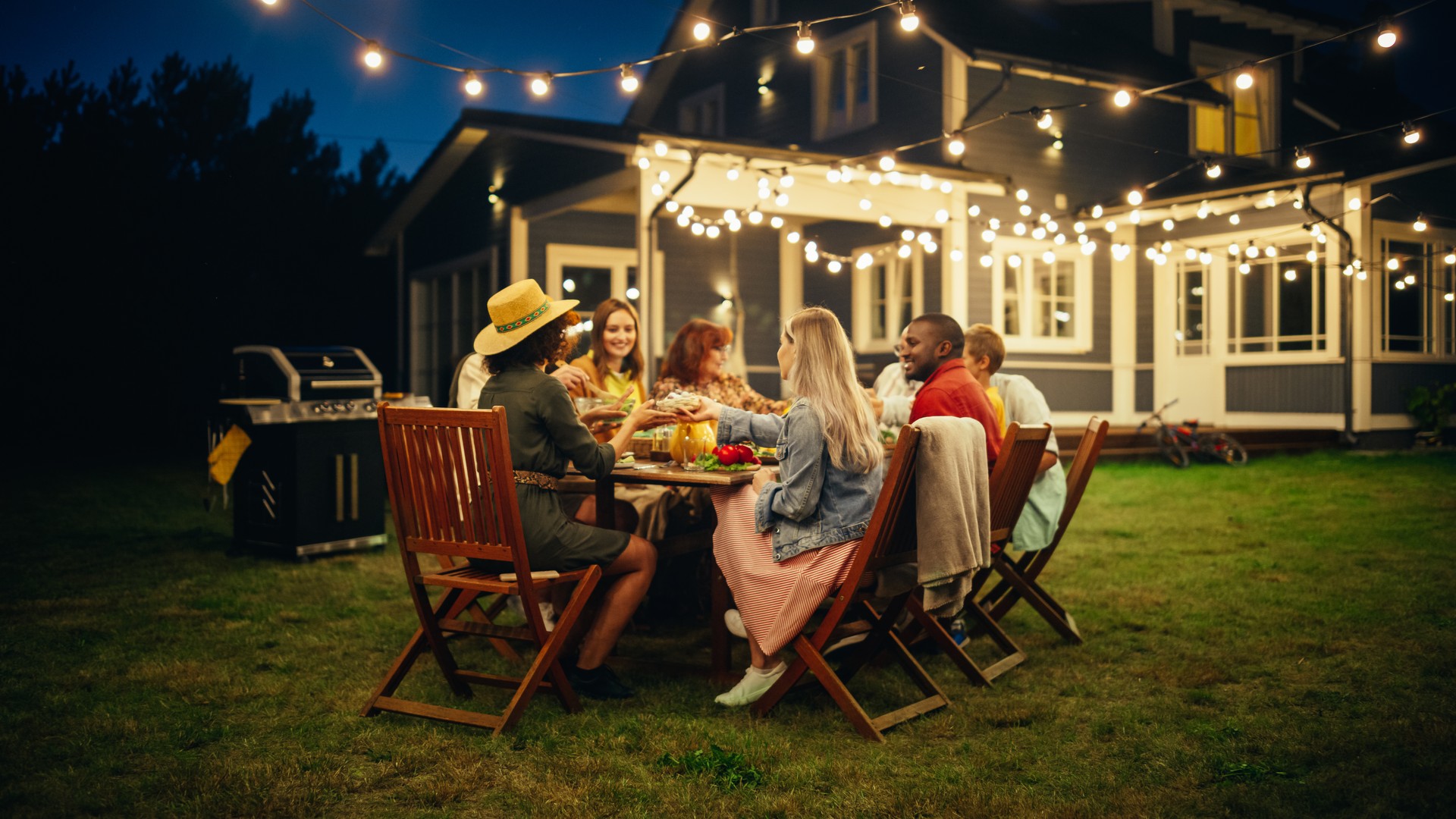 Family and Multicultural Friends Celebrating Outside at Home in the Evening. Group of Children, Adults and Old People Gathered at a Table, Having Fun Conversations. Eating Barbecue and Vegetables.