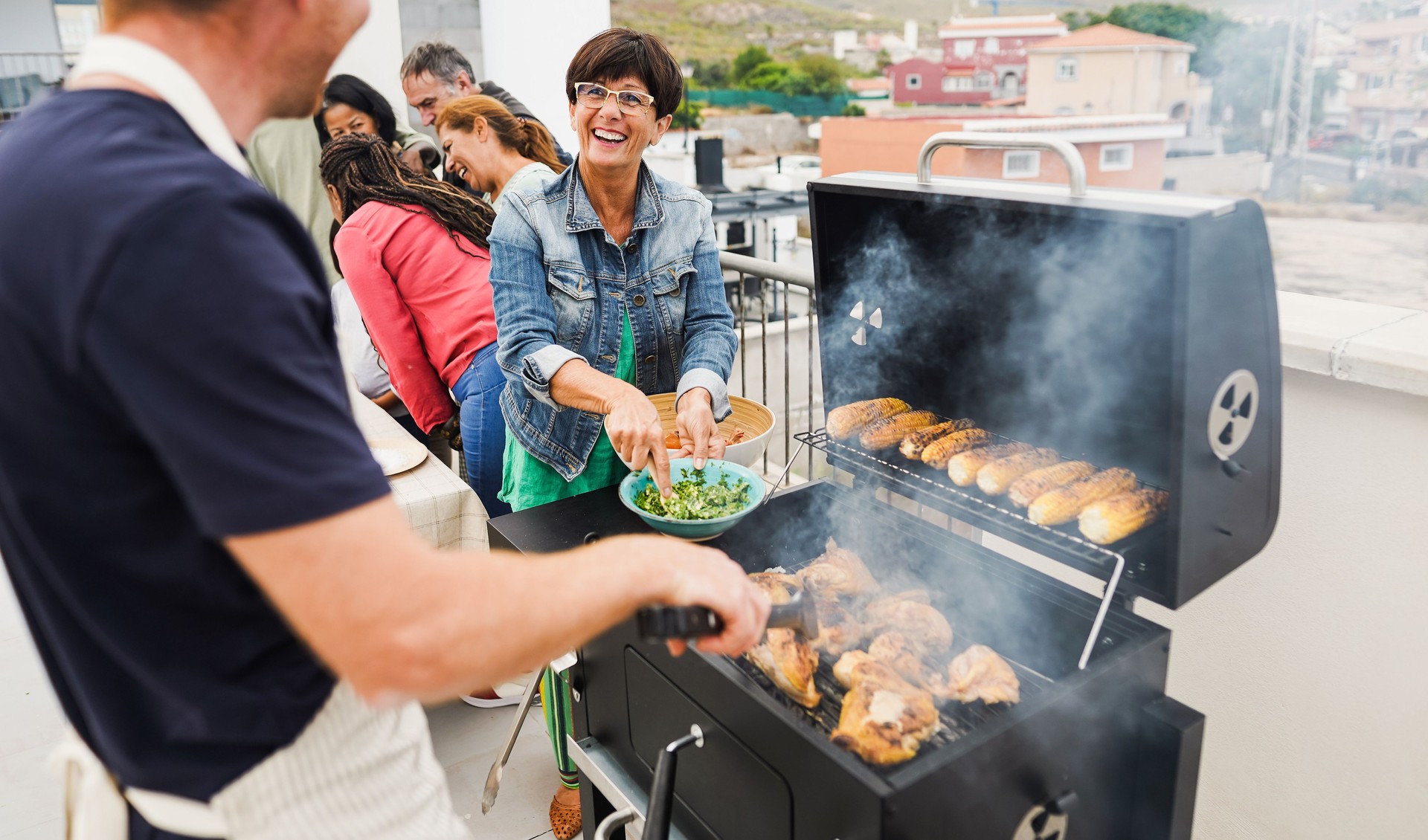 Happy friends cooking corncobs and chicken at barbecue rooftop dinner - Multigenerational people having fun during summer time eating and laughing together - Main focus on woman face